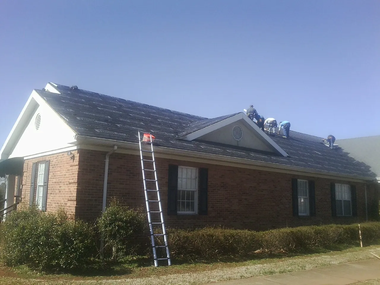 Crew tearing shingles off a brick ranch roof with a ladder set — Metal Roof Repair work by our crew in Splendora