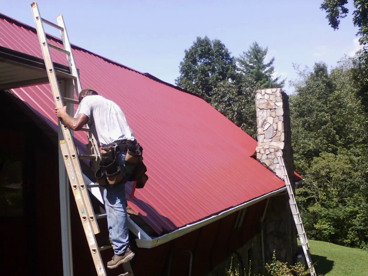 Roofer on a ladder beside a red metal roof and stone chimney — Soffit & Fascia Repair by Roofing Brothers in Splendora